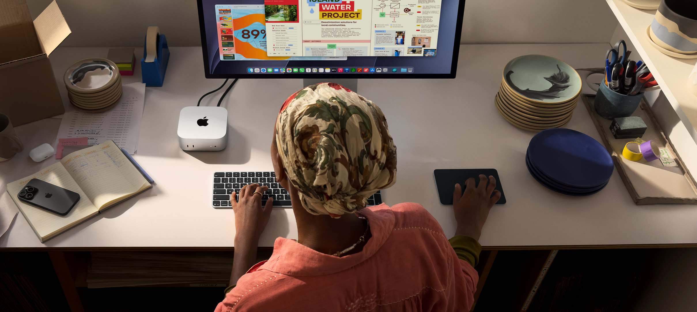 Une personne assise à un bureau utilise un Mac mini avec un Studio Display connecté, Magic Keyboard, Magic Trackpad, divers objets