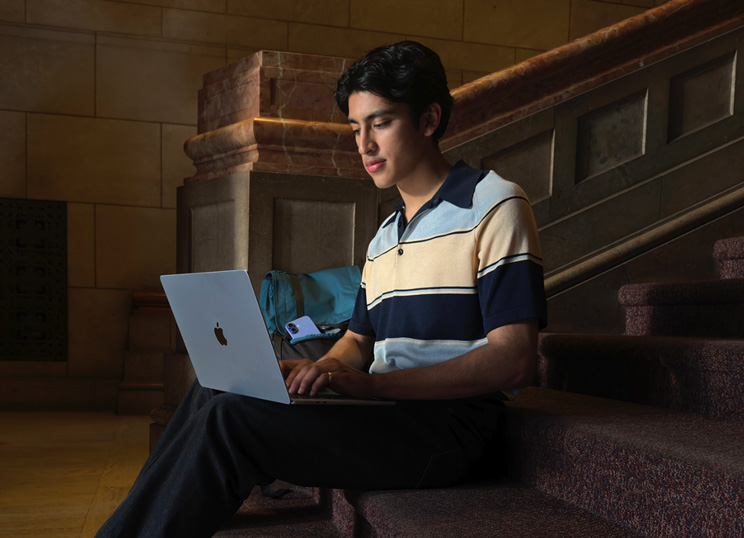 A person sitting on a staircase using a MacBook laptop, focused on work in a warmly lit interior space, with a backpack and iPhone nearby.
