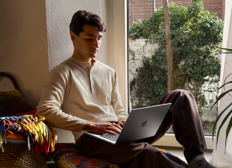 A person working on a Macbook by the window, focused on their screen.