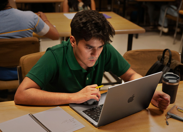 A person working on a Macbook, holding a pencil with notebook and mug on the table.
