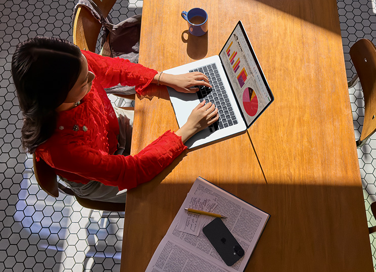 บุคคลหนึ่งใช้ MacBook อยู่บนโต๊ะ โดยมีแก้ว สมาร์ทโฟน และกระดาษอยู่ใกล้ๆ 