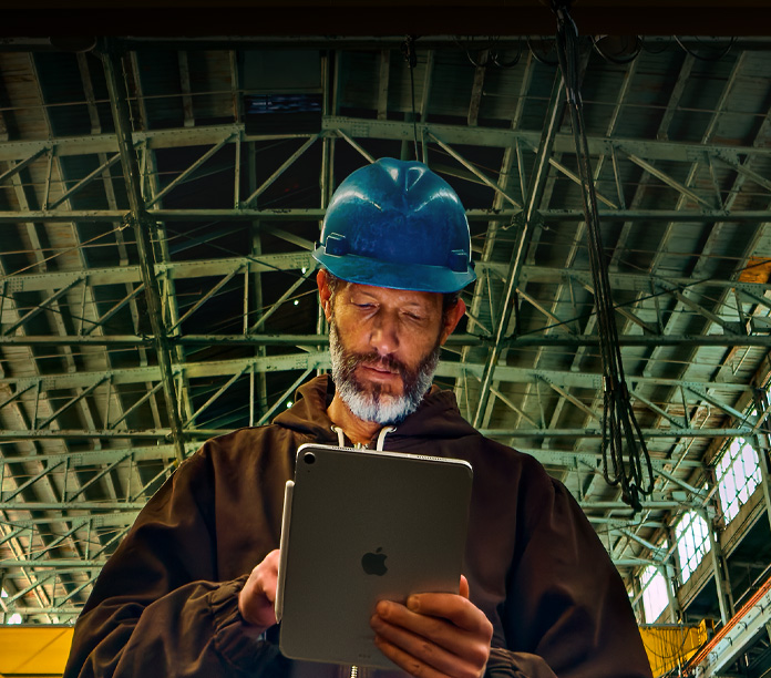 iPad, silver, with Apple Pencil attached, held by a person in a brown jacket and blue safety helmet, working in an industrial setting with metal beams overhead