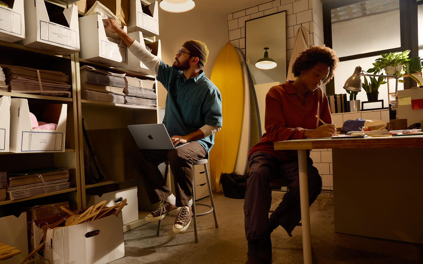 Masada oturan bir kişi kalemle not defterine yazı yazıyor, kucağında MacBook olan diğer bir kişi ise rafların önünde oturmuş, sağ eliyle bir dosyaya uzanıyor.