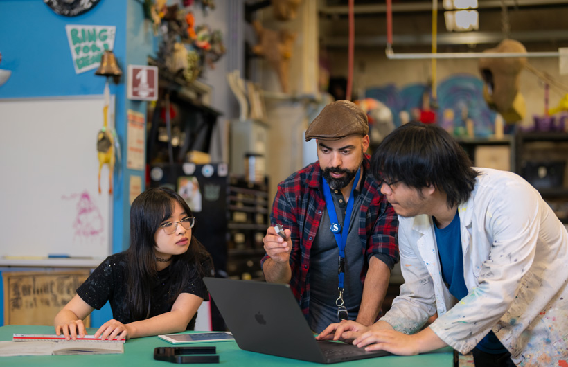 Three students huddle around a MacBook sitting on a table in a cluttered, colourful workshop filled with tools and artwork.