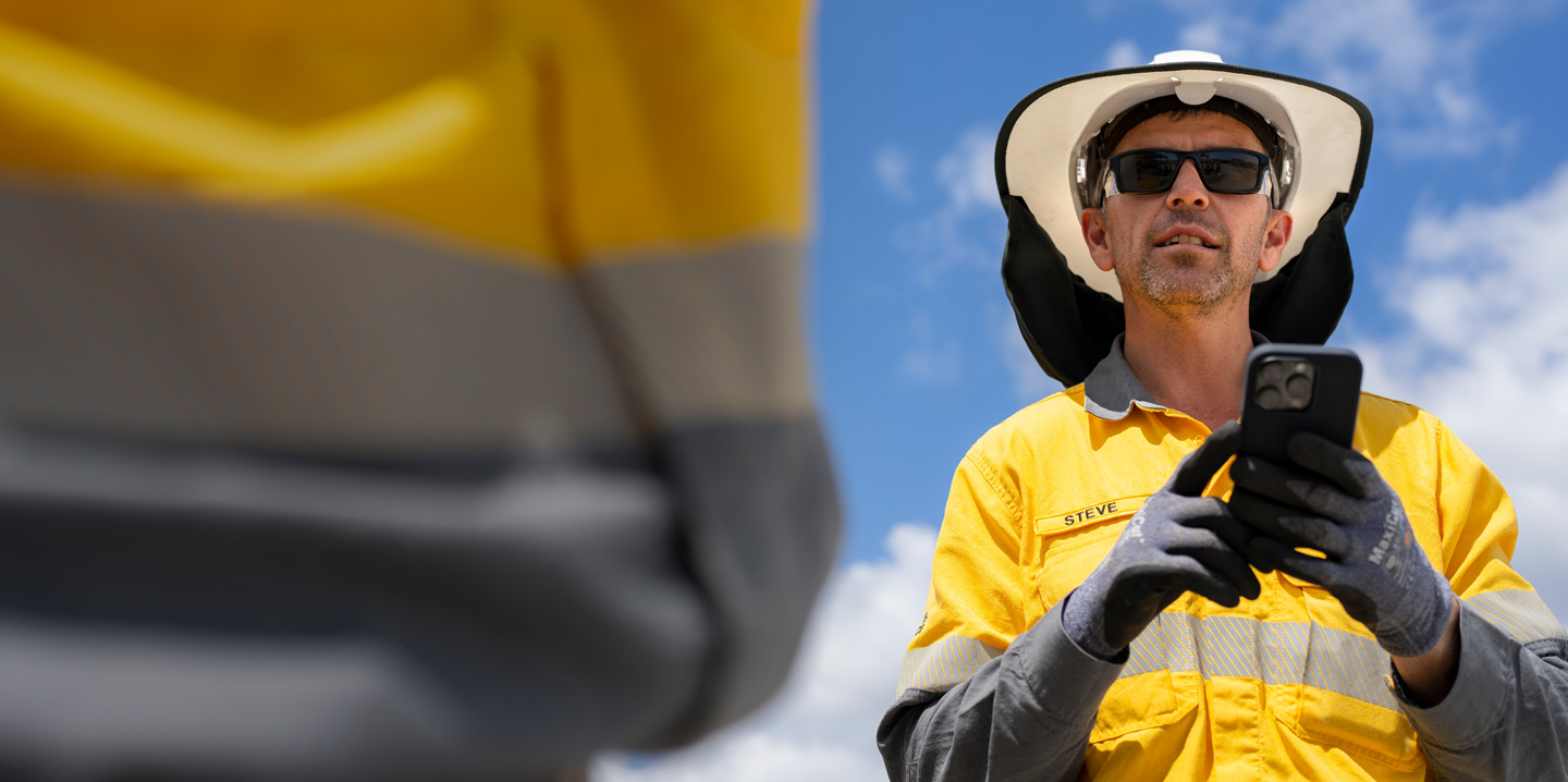 A field worker dressed in safety gear holds an iPhone while talking with a crew member