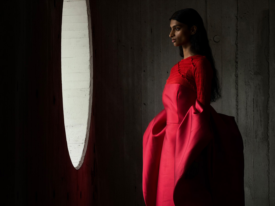 A woman wearing a long red dress stands in an empty room being photographed. The Photo app toggles through the focal lengths to demonstrate the capabilities of the pro camera system, showcasing: 200 mm