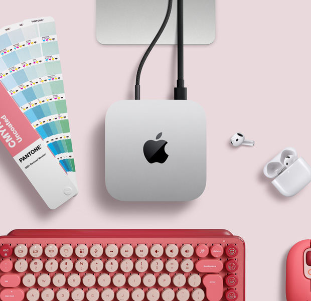 Mac mini, top, silver aluminum, rounded corners, glossy black Apple logo centered, connected by cord, Studio Display stand partially visible, AirPods 4, Pantone color swatches, and red Logitech keyboard and mouse